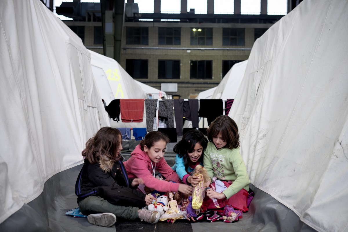On 11 December 2015, girls play with dolls at the refugee shelter at Templehof Airfield in Berlin. (Left-right) sisters Isree, 10,  and Ilaf, 12, from Hassaka, Syria with friends Helen, 10, from Qaamishle, Syria; and Noor, 10, from Idlib, Syria. Templehof Airfield is a former airport dating back to the 1920s, and was used during the Berlin Airlift in 1948. The hangers are today are housing 2,000 refugees, and while designed for transiting within two weeks, many families living there have been there since the shelter opened 6 weeks ago.  Authorities, NGOs and staff working for  a private company running the facility are doing the best they can to make the situation more bearable under quite difficult circumstances. The shelter started operating with two days notice before the first refugees arrived. There are activities for children and they are being scaled up in cooperation with NGOs Save the Children and Together.

"We don't understand the language, and we don't know when we're getting out of there." said Helen, "It's really boring. And at night, it gets really cold. We can't go to the toilets at night, because they're outside and it's freezing cold." Until showers are finished being built on the premises, the residents of Templehof are bussed to nearby public pools once a week to shower.  "The showers are communal and we only have ten minutes to wash," said Helen, "I'm scared the bus taking us back to the camp is going to leave. That happened once and we were stranded [...] We heard a kid was kidnapped and had his throat cut with a knife," she continued, "So we are very afraid. They're just rumours, but we hear a lot of stories about violence, of people being killed [...] One of the good things here are the German classes, and some days circus performers come. They said that they would take us out to a real circus soon. [...] There is a place for kids, but it's been closed for a couple of days now because they're refurnishing it. They're bringing new toys. It's v