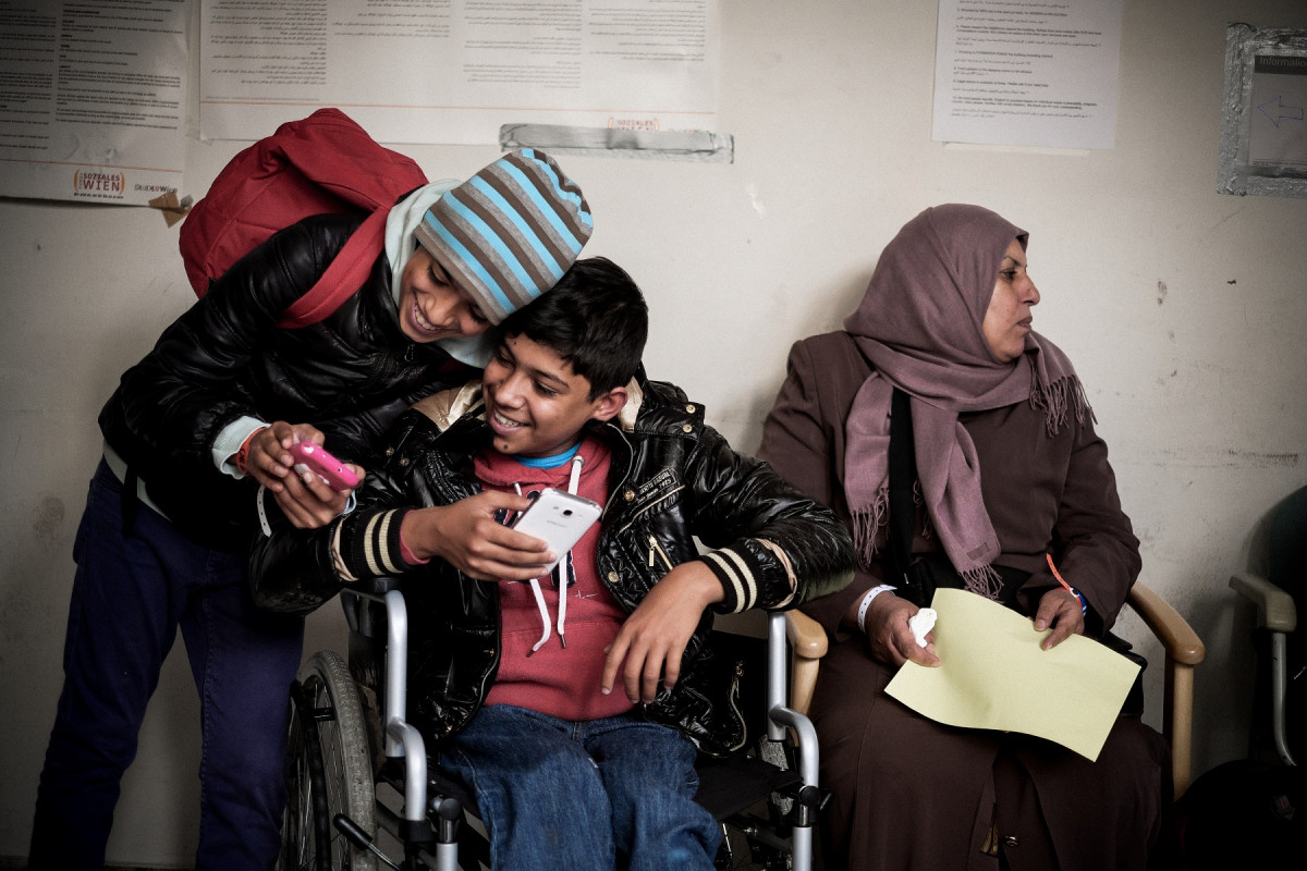 On 4 December 2015 in Austria, 13-year-old Zein Alabdien Al-Faraji and his brother, 15-year-old Sajad Al-Faraji (who uses a wheelchair), laugh while using their cellular phones as they wait with their mother at Kurier Haus, a refugee processing centre in Vienna, the capital, where their family is applying for asylum. The family -- which also includes their sister, Houda Al-Malek – is from Basra Province in Iraq. Refugee and migrant flows into Europe remain at an unprecedented high. Many of them are escaping conflict and insecurity in their home countries of Afghanistan, Iraq, Pakistan and the Syrian Arab Republic.

By late November 2015, refugee and migrant flows into Europe remained at an unprecedented high. Since the beginning of the year, over 870,000 refugees and migrants have crossed the Mediterranean Sea to Europe. Many of them are escaping conflict and insecurity in their home countries of Afghanistan, Iraq, Pakistan and the Syrian Arab Republic. More than one in five is a child. Sajad Al-Faraji, 16, who fled conflict in Basra, Iraq with his brother, Zein Alabdien Al-Faraji, 14, his sister, Houda Al-Malik 26, and his mother Mona Al-Hammoudi, 56, lost the use of his legs during an operation on his lower back when he was one month old in Iraq. His family arrived in Vienna in late 2015 after a harrowing trip through the Balkans.

In November 2015, Houda said “We owe it to my mom that we were able to reach this place.... Since the death of my father, she’s been restlessly struggling to continue with life, giving me and my brothers the motive to continue with our study. And when things deteriorated in Iraq, she decided to get Sajad out of Iraq and bring him here so he would no longer feel inferior.” She added: “My mother took the risk and decided to leave Iraq. She told me she was ready to die in the sea, but the most important thing was that I take my brothers to the country I choose.” Of their sea passage, she said: “We got on that inflatable boa