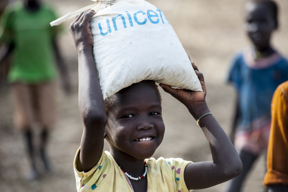 On 18 May, a girl carries atop her head a bag filled with her belongings, in the Bentiu Protection of Civilians site for internally displaced people, in Unity State. The bag bears the UNICEF logo.

By 21 May 2015 in South Sudan, the situation had drastically deteriorated in the preceding two weeks, with heavy fighting ongoing across Unity and Upper Nile States. Some 100,000 people have been internally displaced and 650,000 cut off from aid as humanitarian organizations have been forced to withdraw from affected areas. The violence in the two states has led to a spike in reports of grave child rights violations. Between 13 and 15 May 2015, UNICEF collected testimonies from survivors and witnesses of attacks that took place throughout Rubkona and Guit Counties in Unity State in early May 2015. The testimonies were provided by new arrivals at the Bentiu Protection of Civilians (PoC) site for internally displaced people (IDPs), in Unity State. A total of 41 incidents were recorded, 33 of which are verified. These include 47 children killed, at least 23 children raped and 26 abducted. There are also reports of large-scale recruitment of children. The high numbers of IDPs make it difficult to maintain standards for water, sanitation and hygiene (WASH) services in the Bentiu PoC, where the water supply has fallen from over 15 litres per person per day to 9 litres, and there is only one latrine for every 65 persons. Additionally, several water samples from a new borehole show poor water clarity, meaning water will have to be treated before distribution in order to meet standards in water quality. In the camp, UNICEF is constructing 682 latrines, of which 304 are already complete; supporting partners to provide primary health care; providing measles and polio vaccinations for children under age 15, particularly new arrivals; constructing temporary learning spaces; and, between 8 and 21 May, screened a total of 9,288 children under age 5 for malnutrition. In the past two we